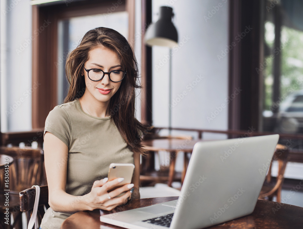 Image of happy woman using laptop and smartphone in cafe. Young beautiful girl sitting in a coffee shop and working on computer Image of happy woman using laptop and smartphone in cafe. Young beautiful girl sitting in a coffee shop and working on computer