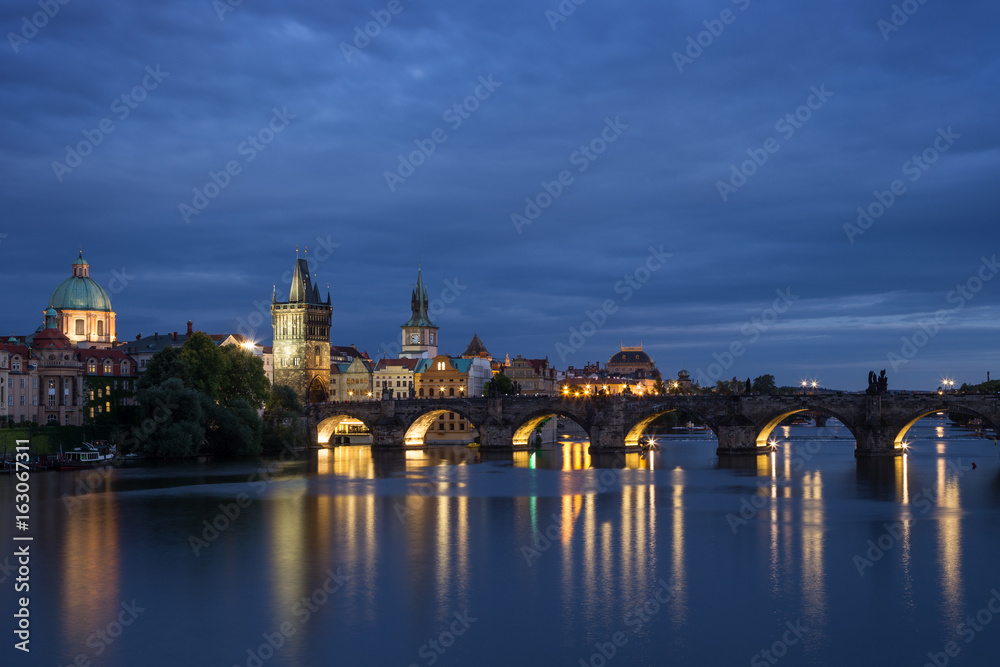 Lit Charles Bridge (Karluv most) and old buildings at the Old Town and their reflections on the Vltava River in Prague, Czech Republic, at dusk. Copy space.