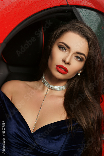 Closeup fashion portrait of young beautiful woman with red lips and long straight brunette hair wearing brilliant shining earrings, necklace choker and blue evening dress sitting in red luxury sport photo