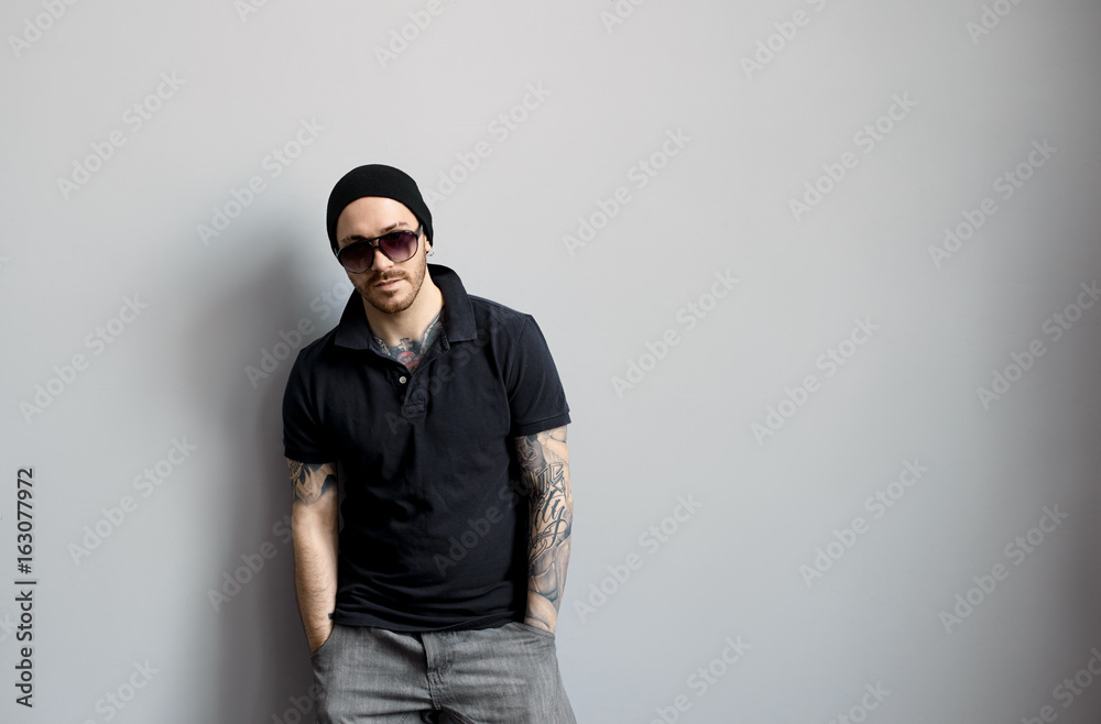 Fototapeta premium Style and fashion concept. Studio shot of trendy handsome bearded young man wearing stylish sunglasses and hat having confident look posing at grey wall, keeping hands in pockets of his jeans