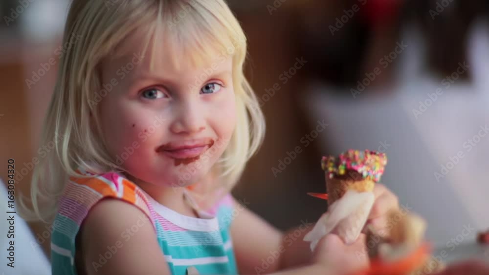 Cute Little Girl Enjoys Her Ice Cream Cone In Ice Cream Shop, She Smiles At Camera Cute Little Girl Enjoys Her Ice Cream Cone In Ice Cream Shop, She Smiles At Camera