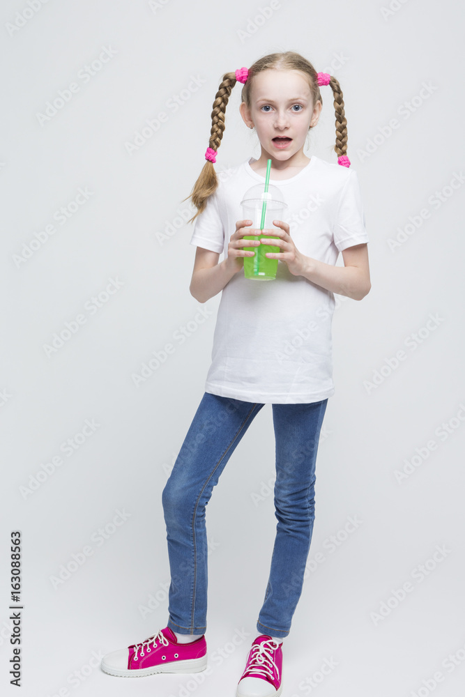 Full Length Portrait of Surprised Pretty Caucasian Blond Girl With Long Pigtails Holding Cup and Drinking Green Juice Through Straw. Full Length Portrait of Surprised Pretty Caucasian Blond Girl With Long Pigtails Holding Cup and Drinking Green Juice Through Straw.