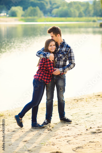 Stylish young smiling couple in shirts and jeans while walking along the lake shore. A beautiful charming girl and her handsome boyfriend leaned against each other.
