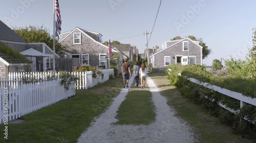 Teens Walk Down Shell Road, Lined With Cottages And American Flags, In New England Seaside 