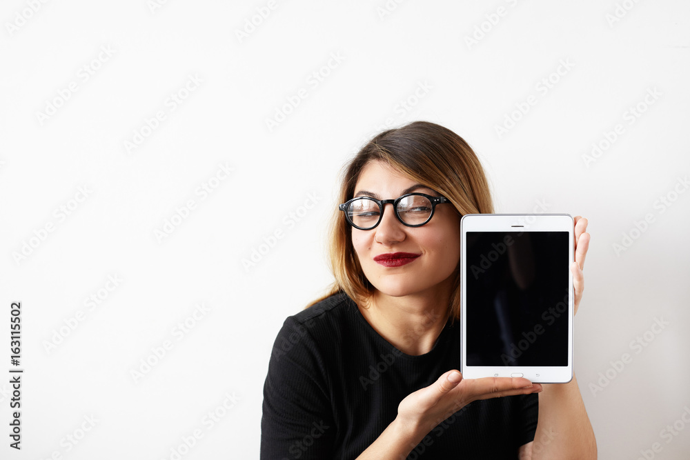 Attractive and confident female entrepreneur in eyewear having cunning look while promoting her product, holding digital tablet with copy space screen for your text or advertising information