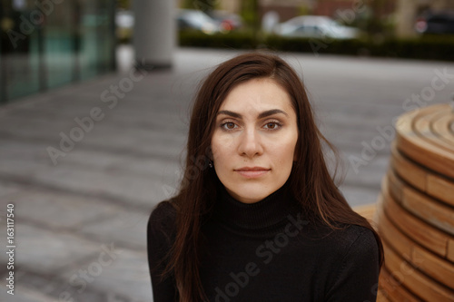 Headshot of attractive confident young woman with long dark hair and brown eyes sitting on bench outdoors, waiting for friends for a walk, looking at camera with serious expression on her face