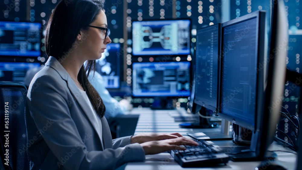 Female Engineer Controller Observes Working of the System. In the Background People Working and Monitors Show Various Information. Female Engineer Controller Observes Working of the System. In the Background People Working and Monitors Show Various Information.