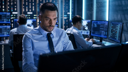 Close-up of Technical Engineer Working on His Computer With Multiple Displays in Monitoring Room. In the Background His Colleagues with Graphics and Various Data on Their Monitors.