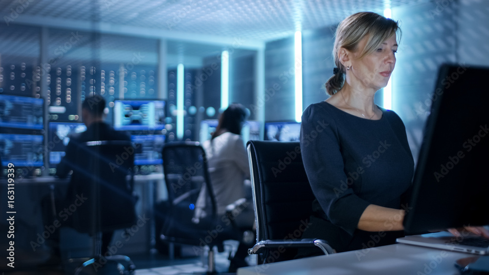 Female IT Engineer Works on Her Desktop Computer in Government Surveillance Agency. In the Background People at Their Workstations with Multiple Screens Showing Graphics. Female IT Engineer Works on Her Desktop Computer in Government Surveillance Agency. In the Background People at Their Workstations with Multiple Screens Showing Graphics.