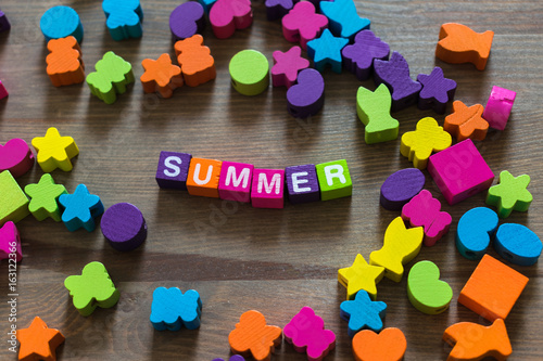 The word Summer made of alphabetic colored wooden cubes on a brown table in an environment of mess from pieces of other games