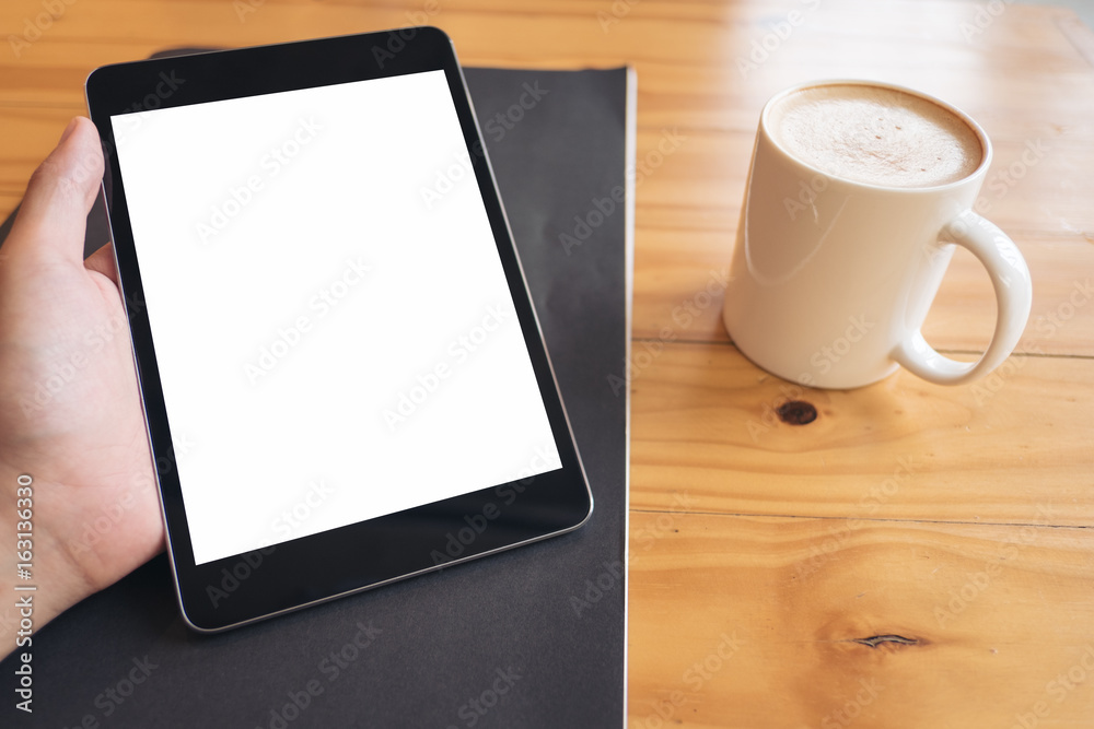 Mock up image of a hand holding black tablet pc with white blank screen and coffee cup on wooden table background in cafe Mock up image of a hand holding black tablet pc with white blank screen and coffee cup on wooden table background in cafe