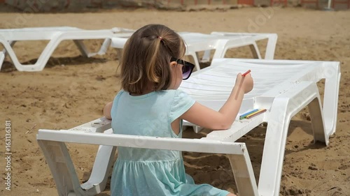 Little girl is sitting on a chaise longue on the beach. She does not get something to draw. Shooting from behind. photo