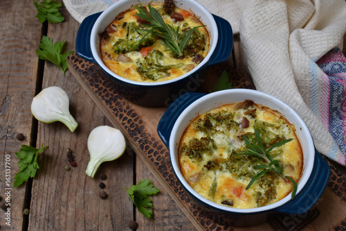 Baked casserole from eggs, bacon, dried tomatoes, broccoli and cheese in blue ramekin on brown wooden background. Mushroom gratin. Breakfast or lanch