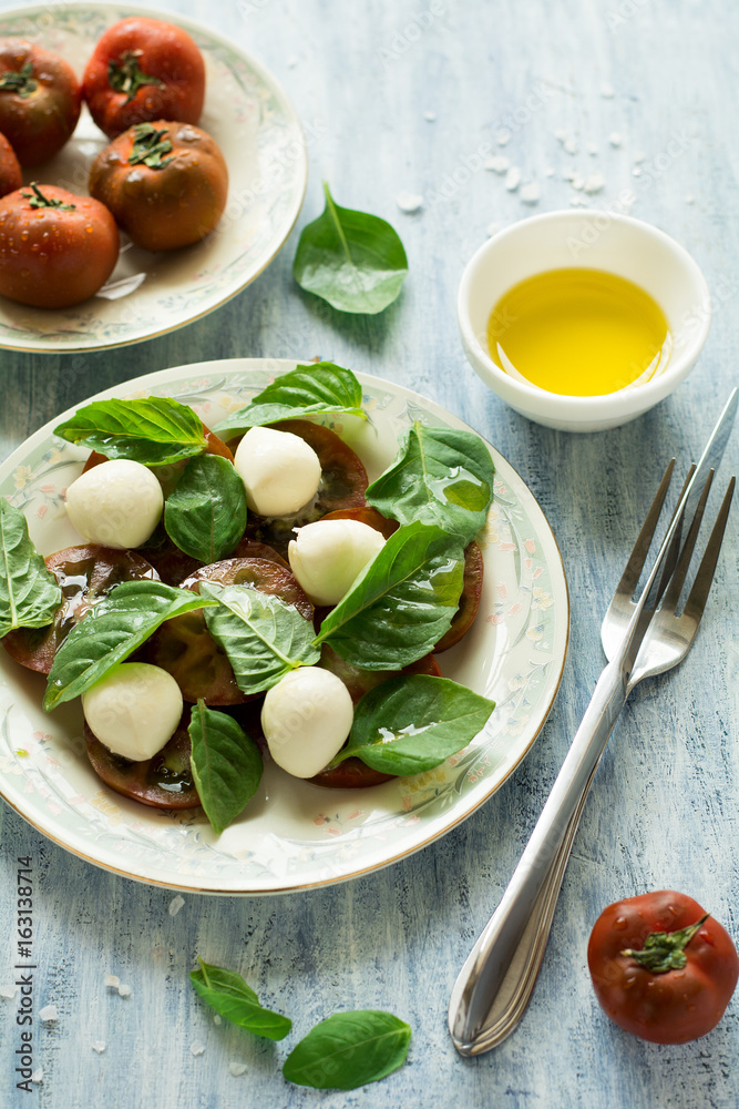Plate of fresh caprese salad with black tomatoes, mini mozzarella and basil leaves on blue wooden table. Selective focus Plate of fresh caprese salad with black tomatoes, mini mozzarella and basil leaves on blue wooden table. Selective focus