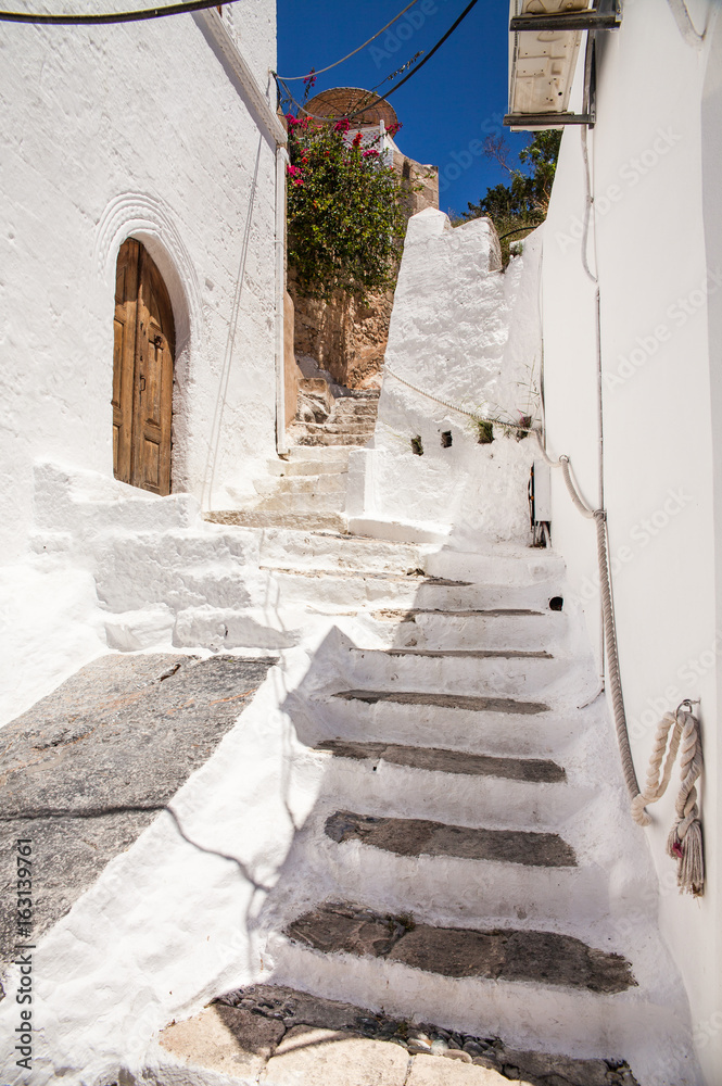 Naklejka premium Narrow streets and typical Greek buildings in the city of Lindos on the island of Rhodes. Residential buildings, shops and restaurants at the foot of the mountain on which the Acropolis is located.