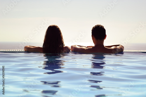 Young Couple Relaxing Together in an Infinity Swimming Pool and Enjoying the View of Horizon and Sea. Travel and Summer Vacation.
