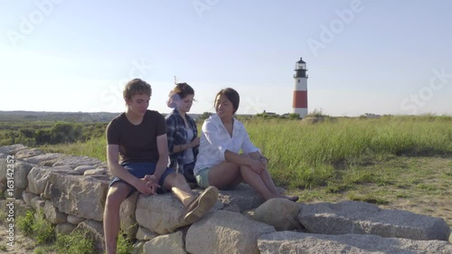 Friends Sit Together On Rock Wall, Lighthouse Behind Them, They Chat And Girl Braids Her Friend's Hair photo