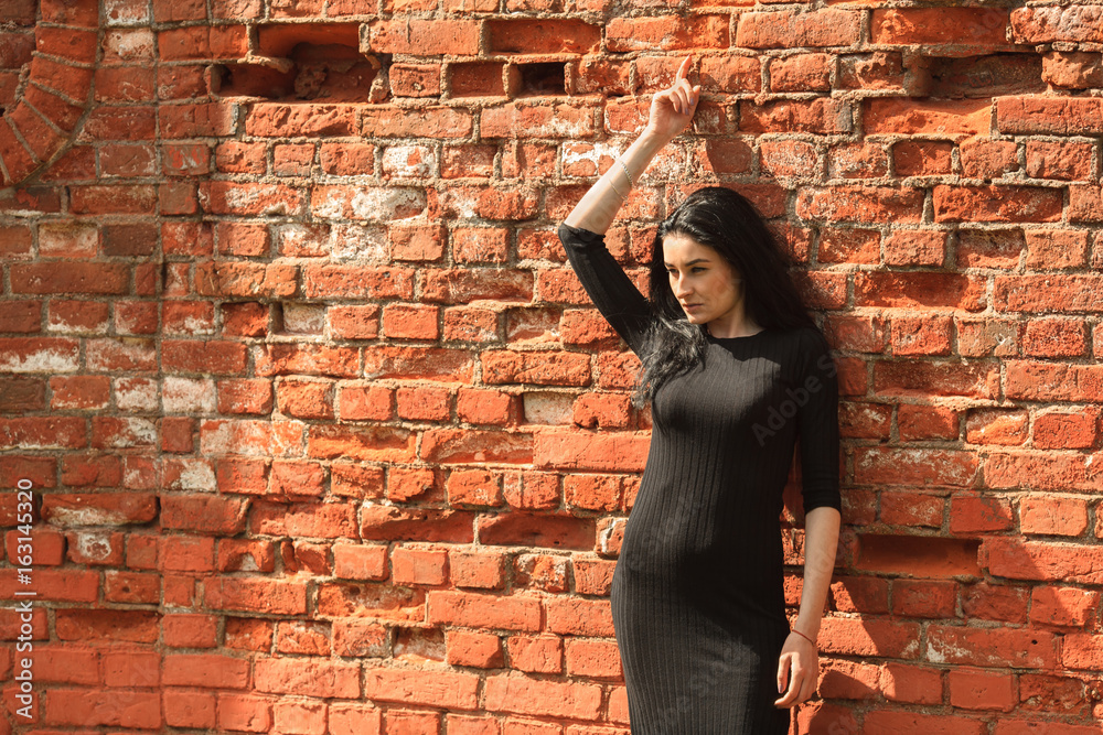 Portrait of young beautiful white girl with aristocratic features with long black hair and in black clothes posing near vintage red brick wall on a summer street.