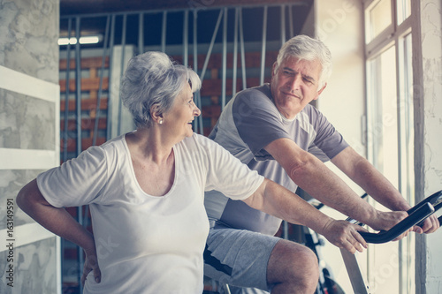 Two senior people at the gym. Senior man sitting on the elliptical machine and having conversation with senior woman.