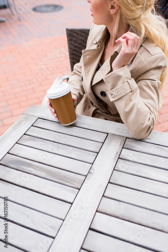 cropped shot of young woman drinking disposable coffee while sitting at cafe on the street