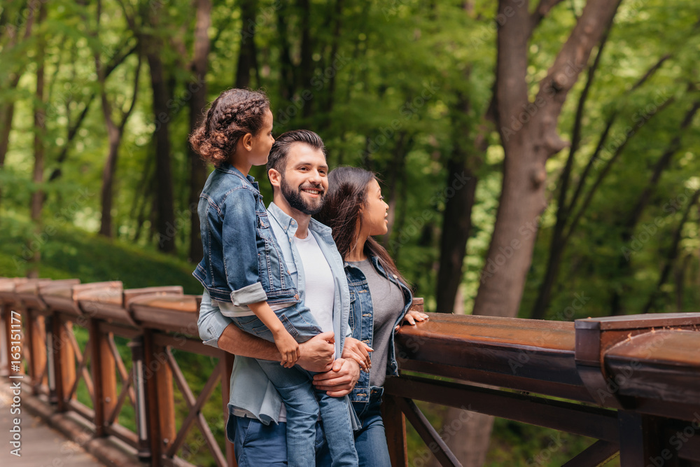 Young happy interracial family standing on wooden bridge, while father holding his daughter in arms Young happy interracial family standing on wooden bridge, while father holding his daughter in arms