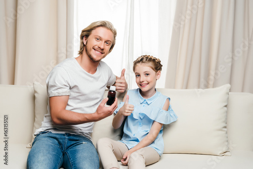 Happy father and daughter holding medicine and showing thumbs up while sitting together on sofa