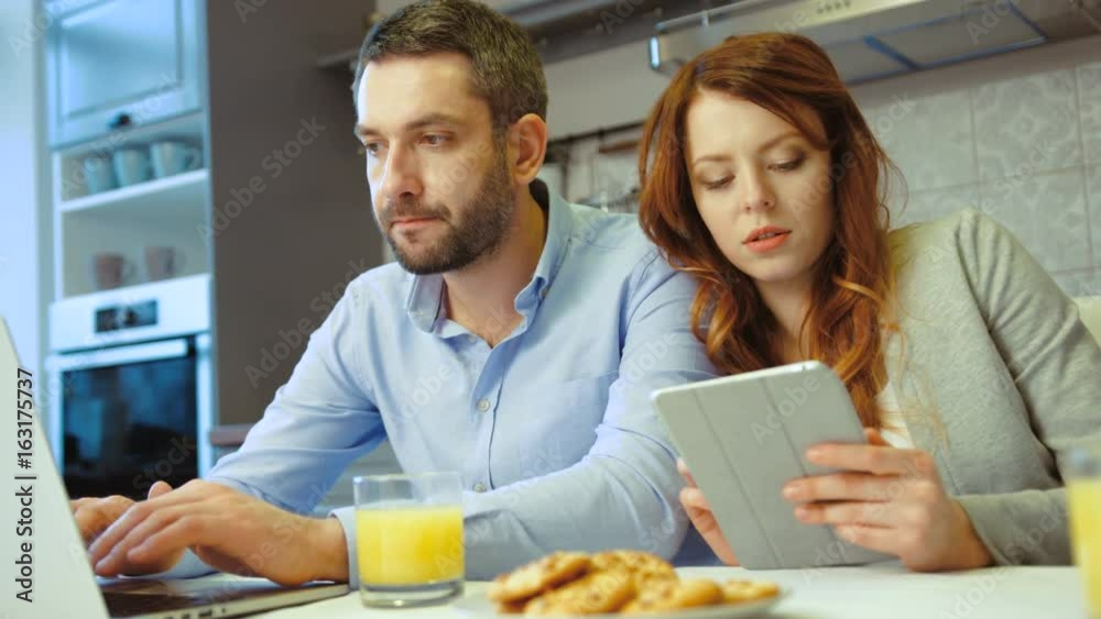 Caucasian young couple siting in kitchen. Young pretty woman using tablet while her husband using laptop. Indoor.