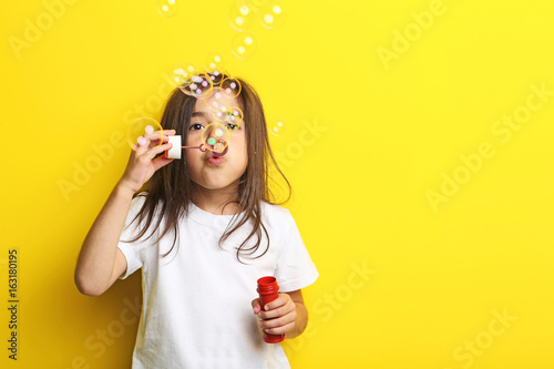 Beautiful little girl blowing soap bubbles on yellow background