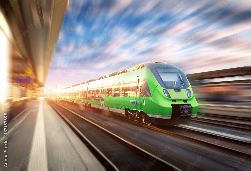 Naklejka premium Szybki pociąg w ruchu na stacji kolejowej o zachodzie słońca w Europie. Piękny zielony nowoczesny pociąg na platformie kolejowej z efektem rozmycia ruchu. Przemysłowa scena z pociągiem pasażerskim na linii kolejowej