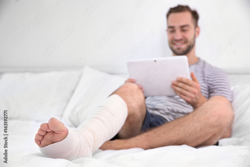 Handsome young man with tablet computer and bandaged leg sitting on bed at home Handsome young man with tablet computer and bandaged leg sitting on bed at home