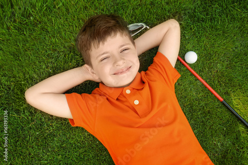 Cute boy with ball and driver lying on golf course