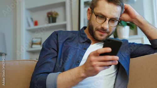 Young caucasian man with glasses and beard relaxing on couch using smart phone in modern industrial house. Indoor.