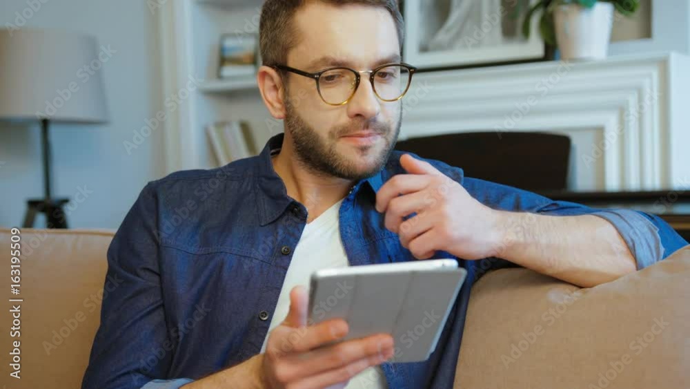 Concentrated caucasian man with glasses and beard siting on sofa using tablet in living room. Indoor.