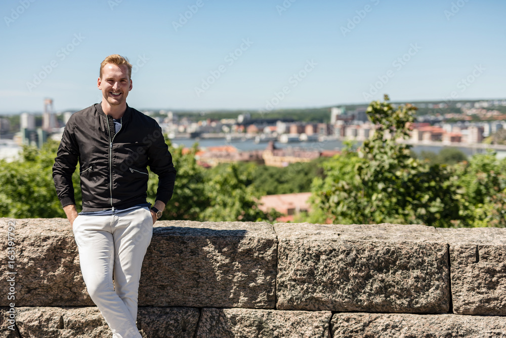 Trendy man in black jacket standing high up looking at the view of the city, cityscape behind him on a sunny summer day. Trendy man in black jacket standing high up looking at the view of the city, cityscape behind him on a sunny summer day.