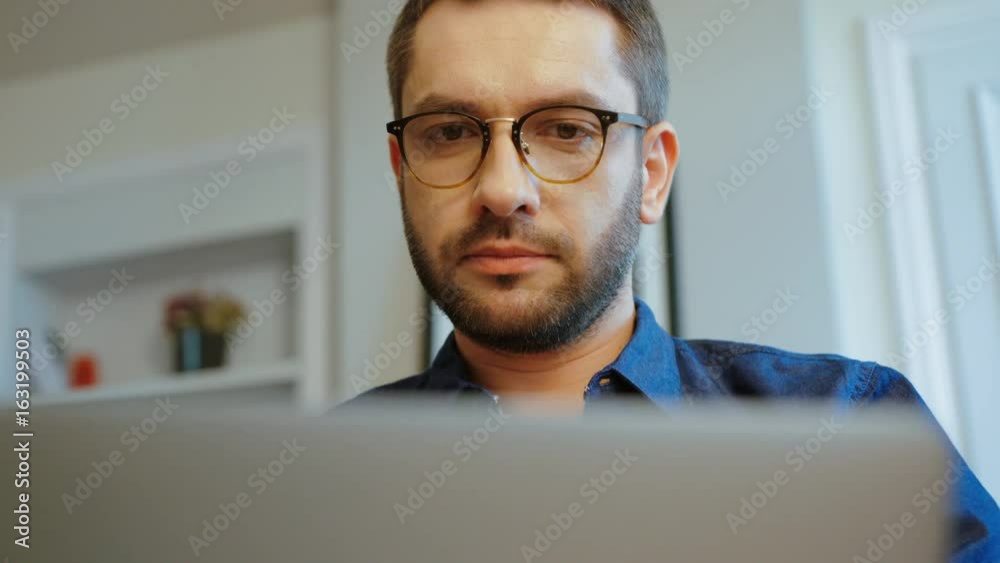 Portrait of serious caucasian young man in glasses with beard in blue shirt using laptop in modern office. Indoor.