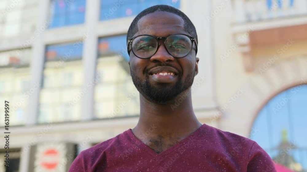 Portrait of handsome african american man in glasses and T-shirt smiling on city background. Outdoor. Portrait of handsome african american man in glasses and T-shirt smiling on city background. Outdoor.