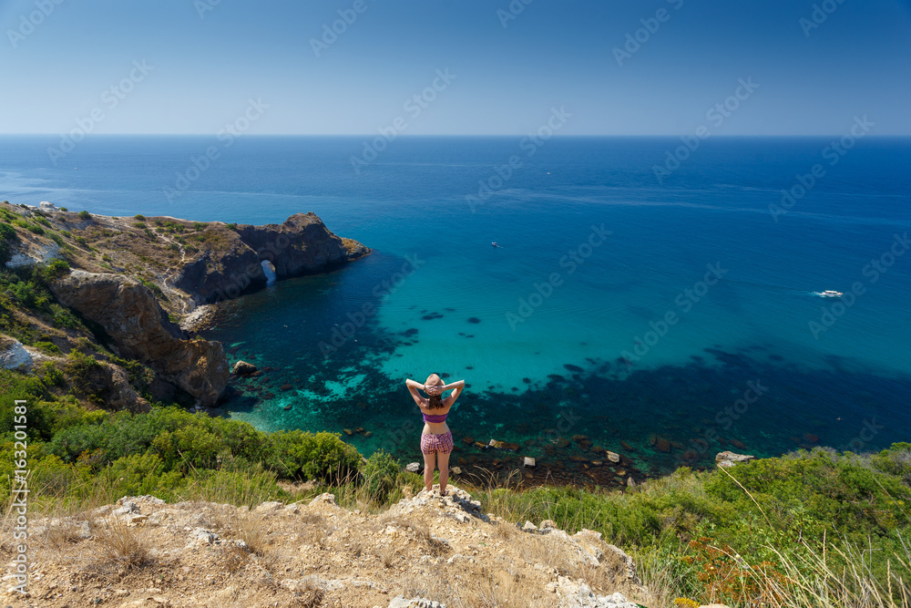 Obraz premium Beautiful woman standing on a rock above the Black sea and looking at calm and deep blue water. Feel free to explore, adventure and discovery nature treasure of nature.From the back. Top view.