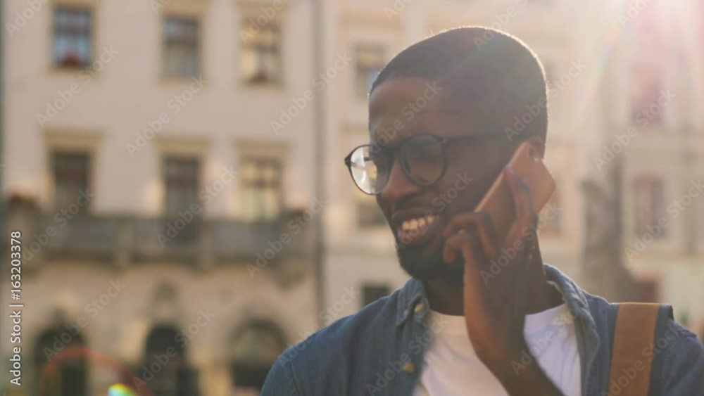 Portrait of young african man in blue shirt talking on mobile phone with friend and smiling with teeth in city. Outdoor. Portrait of young african man in blue shirt talking on mobile phone with friend and smiling with teeth in city. Outdoor.