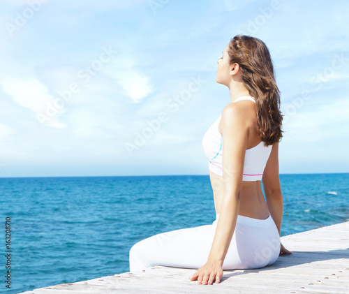Attractive, beautiful and young girl in white sportswear meditating on a wooden pier at summer. Yoga, sport, recreation, vacation and traveling concept. © Acronym