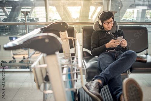 Young asian man using smartphone and listening to music while waiting for connecting flight on bench in the international airport terminal, travel abroad concept