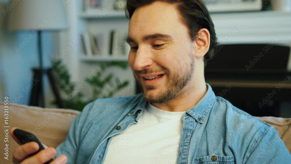 Portrait of attractive man in casual shirt typing on the smart phone, sitting on the sofa at the living room. Portrait of attractive man in casual shirt typing on the smart phone, sitting on the sofa at the living room.