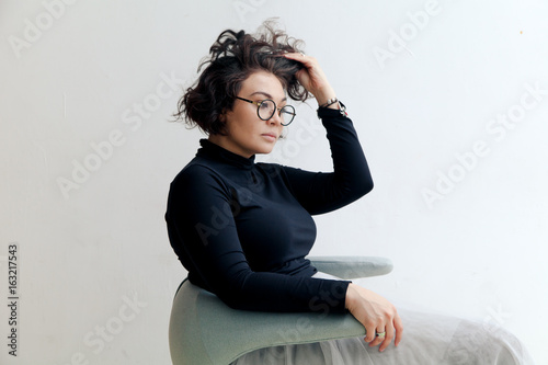 Young plus size female model in tutu skirt sitting in chair stretching her dark curvy hair in bright empty room. Composition with copy space photo