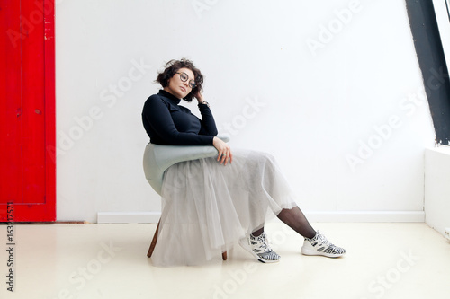 Young plus size female model in tutu skirt sitting in chair stretching her dark curvy hair in bright empty room. Composition with copy space