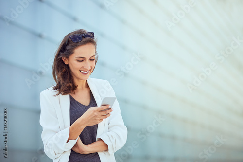 Laughing young businesswoman texting coworker in bright modern outside setting