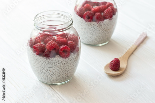 Chia seeds pudding with coconut milk and raspberry in glass jars. Wooden spoon with berry in white wooden table.