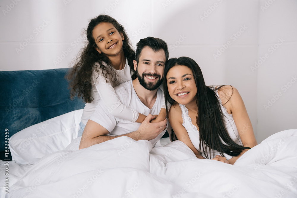 Happy multiethnic family with one child hugging and smiling at camera in bedroom Happy multiethnic family with one child hugging and smiling at camera in bedroom