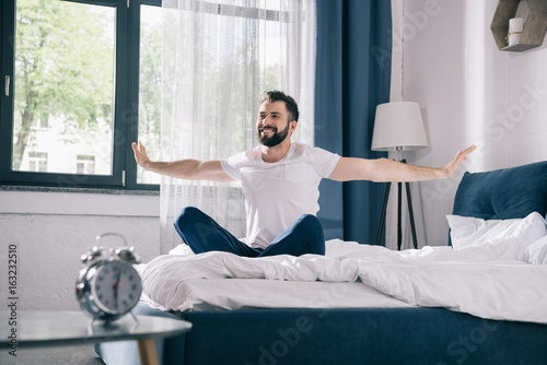 Smiling young man in pajamas stretching while sitting on bed at morning