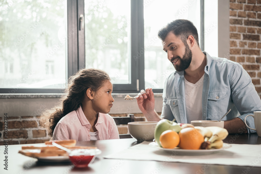 portrait of father feeding grumpy daughter with porridge during breakfast portrait of father feeding grumpy daughter with porridge during breakfast