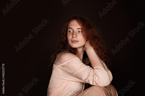 attractive redhead woman sitting in beige clothes, isolated on brown