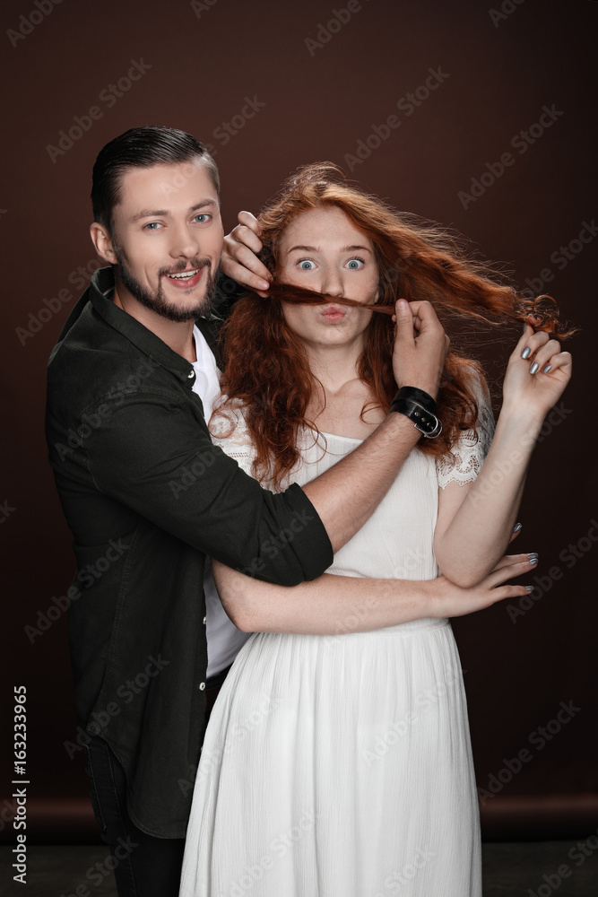 Young cheerful couple, man making mustache of hair for his girlfriend, isolated on brown Young cheerful couple, man making mustache of hair for his girlfriend, isolated on brown
